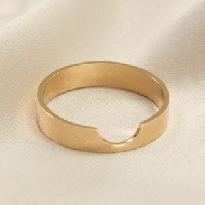 May include: A gold-colored ring with a unique cutout design. The ring is made of a solid metal and has a smooth, matte finish. The ring is set against a cream-colored fabric background.