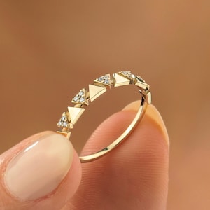 May include: A gold ring featuring a series of small, triangular shapes. Each triangle is accented with small, clear gemstones. The ring is held between two fingers against a blurred background.