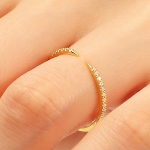 May include: A gold-coloured open ring with small, clear gemstones set along the band. The ring has a split design, with the ends curving away from each other. The ring is worn on a finger, with the skin visible in the background.