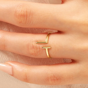 May include: A gold-colored ring with a modern, minimalist design. The ring features a horizontal bar and a vertical bar, creating a unique open design. The ring is worn on a finger, showcasing its sleek and contemporary style.