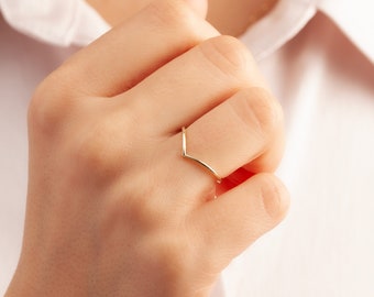 May include: A thin, polished gold ring featuring a V-shaped chevron design. The band has a smooth, metallic finish and a pointed centre point that creates a distinct angular silhouette when worn on the finger, showcasing a simple, refined aesthetic.