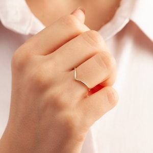 May include: A gold-colored ring with a V-shaped design. The ring is worn on the middle finger of a person's hand.