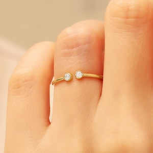 May include: A gold-colored ring with two small, clear gemstones set on either end. The ring is open at the front, creating a unique design. The ring is worn on a finger.