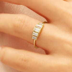 May include: A gold ring featuring a row of five rectangular, clear gemstones. The ring is set on a slender gold band. The gemstones are aligned horizontally, creating a linear design. The ring is shown on a person's finger.