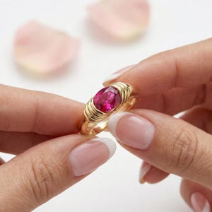 May include: A gold ring with a large, oval ruby gemstone. The ring's band has horizontal gold ridges, giving a textured look. The ruby is a bright red, set in a classic, elegant design. A beautiful piece of jewellery.