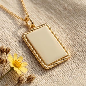 May include: A gold-colored rectangular pendant necklace with a cream-colored center. The pendant has a rope-like border and hangs from a delicate chain. A small yellow flower and dried seed pods are in the lower left corner.
