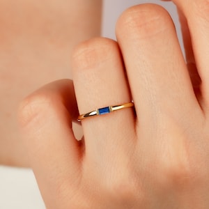 May include: A thin gold band ring with a small, rectangular blue sapphire gemstone.