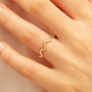 May include: A gold-colored ring with a wavy design. The ring has a textured surface and is worn on a finger. The ring's design resembles a zig-zag pattern. The ring is made of a shiny, metallic material.