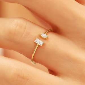 May include: A gold-colored ring with two clear, rectangular and oval-shaped gemstones. The ring has a split shank design, with one gemstone on each side of the split. The ring is shown on a person's finger.