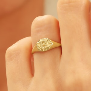 Könnte beinhalten: Ein goldfarbener Ring mit dem Buchstaben „B“ in der Mitte, umgeben von einem Sonnenstrahl-Design. Der Ring wird an einem Finger getragen und zeigt seine polierte Oberfläche und die filigranen Details. Ein Schmuckstück.