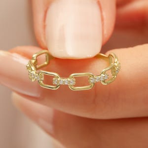 May include: A gold-coloured chain link ring with small, clear gemstones set into the links. The ring is held by a person with a light skin tone. The ring is a delicate piece of jewellery.