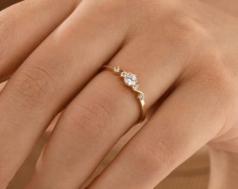 May include: A delicate gold ring with a small round diamond in the center and two smaller diamonds on either side. The ring is set on a curved band.