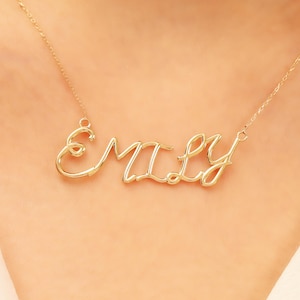 May include: A gold-colored necklace with the name "EMILY" in a cursive font. The necklace is suspended from a delicate chain. The name is the focal point, crafted in a stylish, flowing script.