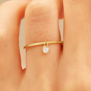 May include: A delicate gold ring with a small, dangling, round, clear gemstone. The ring is thin and simple, with a minimalist design. The gemstone is set in a gold bezel and hangs freely from the ring's band.