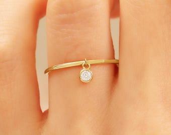 May include: A delicate gold ring with a small, dangling, round, clear gemstone. The ring is thin and simple, with a minimalist design. The gemstone is set in a gold bezel and hangs freely from the ring's band.