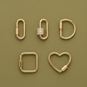 May include: A collection of five gold-toned metal carabiner clasps in various shapes including oval, D-shape, square, and heart. One oval clasp features a cylindrical accent embedded with small clear stones. Each clasp has a spring-loaded gate.