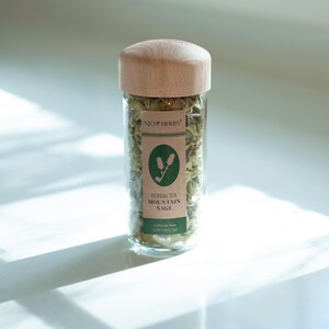May include: A glass jar with a wooden lid filled with dried mountain sage herbs. The label reads "NJOY HERBS" and "HERBAL TEA MOUNTAIN SAGE".