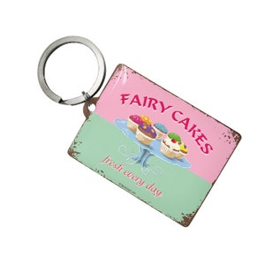 May include: A pink and green metal keychain with a vintage design. It features a sign that says "Fairy Cakes" with a picture of cupcakes on a cake stand. The text below the sign says "fresh every day".