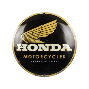 Può includere: Un cartello in metallo nero e oro con il logo Honda Motorcycles. Il logo presenta un'ala stilizzata e il testo "HONDA MOTORCYCLES HAMAMATSU, GIAPPONE".
