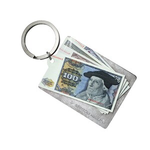 Könnte beinhalten: Ein silberner Schlüsselanhänger mit einer Nachbildung eines 100 Deutsche Mark-Scheins, auf dem ein Porträt eines Mannes mit einem schwarzen Hut zu sehen ist. Der Text "Banknote" und "100 Deutsche Mark" ist auf dem Schein sichtbar.