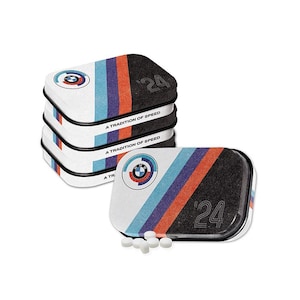 May include: A stack of four white and black mint tins with a blue, red and purple stripe design. The tins feature the BMW logo and the text "A Tradition of Speed" and "'24".  A scattering of white mints are in front of the tins.