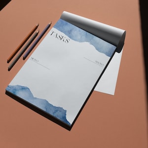 May include: A white notepad with the word "TASKS" printed at the top. The notepad has a blue watercolour design at the bottom and is lying on a light brown surface. Three coloured pencils are lying next to the notepad.