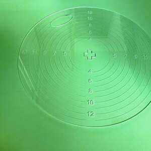 May include: A transparent circular target with concentric rings and numbers from 4 to 13. The target features a crosshair at the centre and a small handle hole at the top. The target is made of clear material.