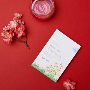 May include: A wedding invitation with the names "MARY JANE and PETER PARKER" is displayed on a red surface. The invitation features a watercolor design of flowers and grass. A glass of pink liquid and pink flowers are also present.