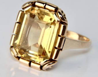 Pode incluir: Um anel de ouro com uma grande gema amarela clara e transparente engastada em um engaste de bezel. O anel tem uma banda decorativa com um padrão repetitivo.