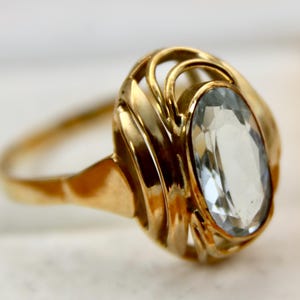 Puede incluir: Un anillo de oro con una gran piedra preciosa ovalada transparente engastada en un diseño delicado e intrincado.
