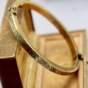 Puede incluir: Un brazalete de oro con un intrincado diseño floral grabado en la superficie. El brazalete se muestra en una caja de madera.