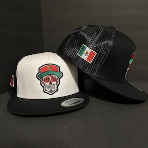 May include: Two snapback hats. One is black with a mesh back and a Mexican flag patch. The other is white and black with a sugar skull design and a Mexican flag patch. Both hats have a flat brim.