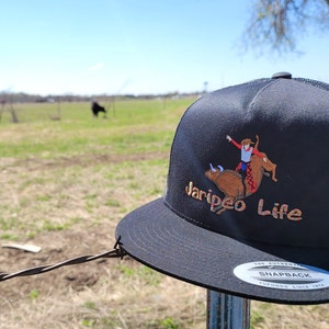 May include: Black trucker hat with a mesh back and a flat brim. The hat features an embroidered design of a rodeo rider on a bull, with the words "Jaripeo Life" in a stylized font. The hat is displayed outdoors.