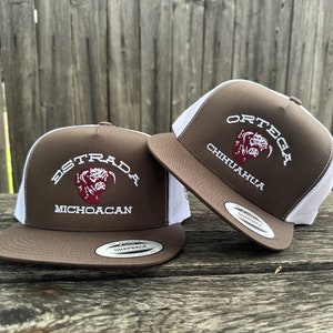 May include: Two brown and white trucker hats with embroidered text. One hat reads "ESTRADA MICHIOACAN" and the other reads "ORTEGA CHIHUAHUA". Both hats have a brown brim and a white mesh back.