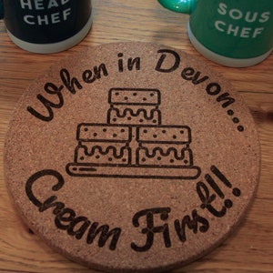 May include: A round cork coaster with the text "When in Devon... Cream First!!" and a graphic of a stack of cream teas. The coaster is brown with a light brown border.