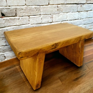 May include: A rustic wooden stool with a natural wood finish. The stool has a rectangular top and four legs. The stool is made of solid wood and has a natural wood grain.
