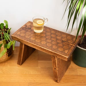May include: A wooden stool with a honeycomb pattern on the top. The stool is stained a dark brown colour and has a rustic look. A glass of tea sits on top of the stool.