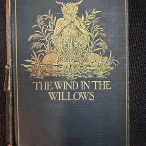 Pode incluir: Livro antigo de capa dura intitulado "O Vento nos Salgueiros". A capa é azul escuro com letras douradas e uma ilustração dourada de uma figura mítica tocando uma flauta, rodeada por plantas e animais.