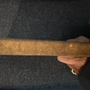 May include: A rectangular, wooden object with a textured, light brown surface. The object is held in a hand, with a dark background. The item appears to be a tool or part of a larger item.