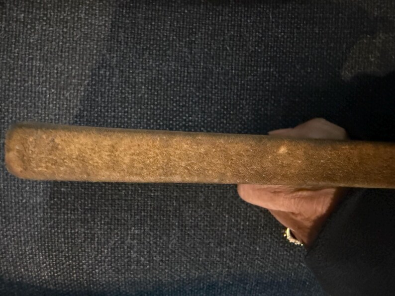May include: A rectangular, wooden object with a textured, light brown surface. The object is held in a hand, with a dark background. The item appears to be a tool or part of a larger item.