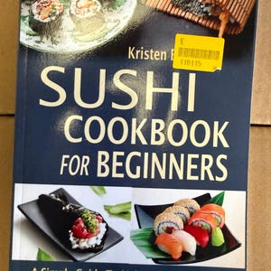 Op de afbeelding: Een kookboek getiteld Sushi Cookbook for Beginners met een donkerblauwe kaft met afbeeldingen van diverse sushi-rolletjes, een bamboe rolmatje en de ondertitel A Simple Guide To Making Sushi At Home With Over 70 Delicious Sushi Recipes.
