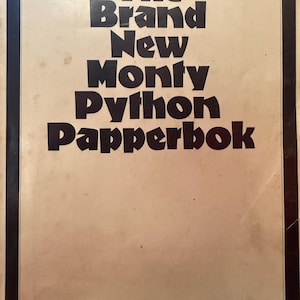May include: A rectangular book cover with a cream-coloured background and a thick black border. The text "The Brand New Monty Python Papperbok" is printed in large, bold, black, sans-serif capital letters centred on the front cover of the book.