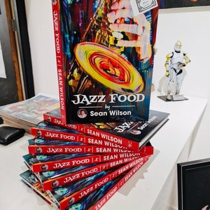May include: A stack of red and white paperback books titled "Jazz Food" by Sean Wilson. The cover of the top book features a colorful illustration of a saxophone.