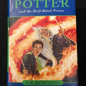 Puede incluir: Un libro de tapa dura azul titulado "Harry Potter y el Misterio del Príncipe". La portada muestra a Harry Potter, un mago y llamas. El nombre de la autora, J.K. Rowling, y la editorial, Bloomsbury, también son visibles.