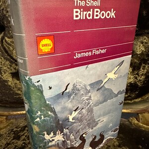 Könnte beinhalten: Ein rotes und weißes Buch mit dem Titel "The Shell Bird Book" von James Fisher. Der Buchumschlag zeigt ein Shell-Logo und eine farbenfrohe Illustration von Vögeln auf einer Klippe.