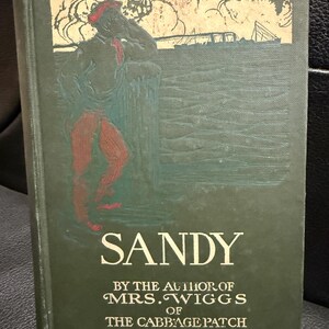 May include: Antique book titled "Sandy" by Mrs. Wiggs of the Cabbage Patch. The cover is dark green with an illustration of a person in red pants and a red hat. The title is in large white letters.