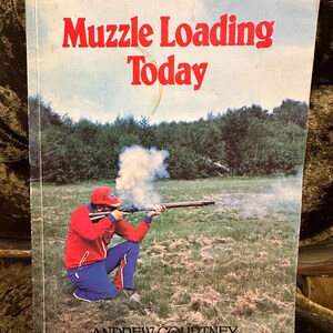 Könnte beinhalten: Ein Buch mit dem Titel "Muzzle Loading Today" von Andrew Courtney. Der Umschlag zeigt eine Person in einer roten Jacke und blauen Hosen, die in einem grasbewachsenen Feld ein Vorderladergewehr abfeuert.