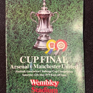 May include: A vintage souvenir program cover for the 1979 FA Cup Final, featuring a silver trophy atop a green grass background. The text reads "CUP FINAL Arsenal v Manchester United" with "Wembley Stadium" in red.