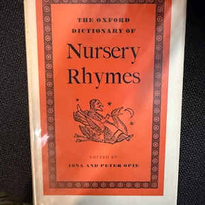 Puede incluir: Un libro antiguo titulado "The Oxford Dictionary of Nursery Rhymes". La portada naranja vibrante presenta texto negro y bordes decorativos. Se presenta una ilustración de una figura montada en un pájaro. Editado por Iona y Peter Opie.