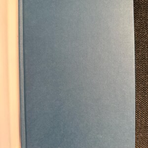 May include: A hardcover book with a light blue cover. The book is open, revealing the white pages on the left side. The cover has a textured appearance.